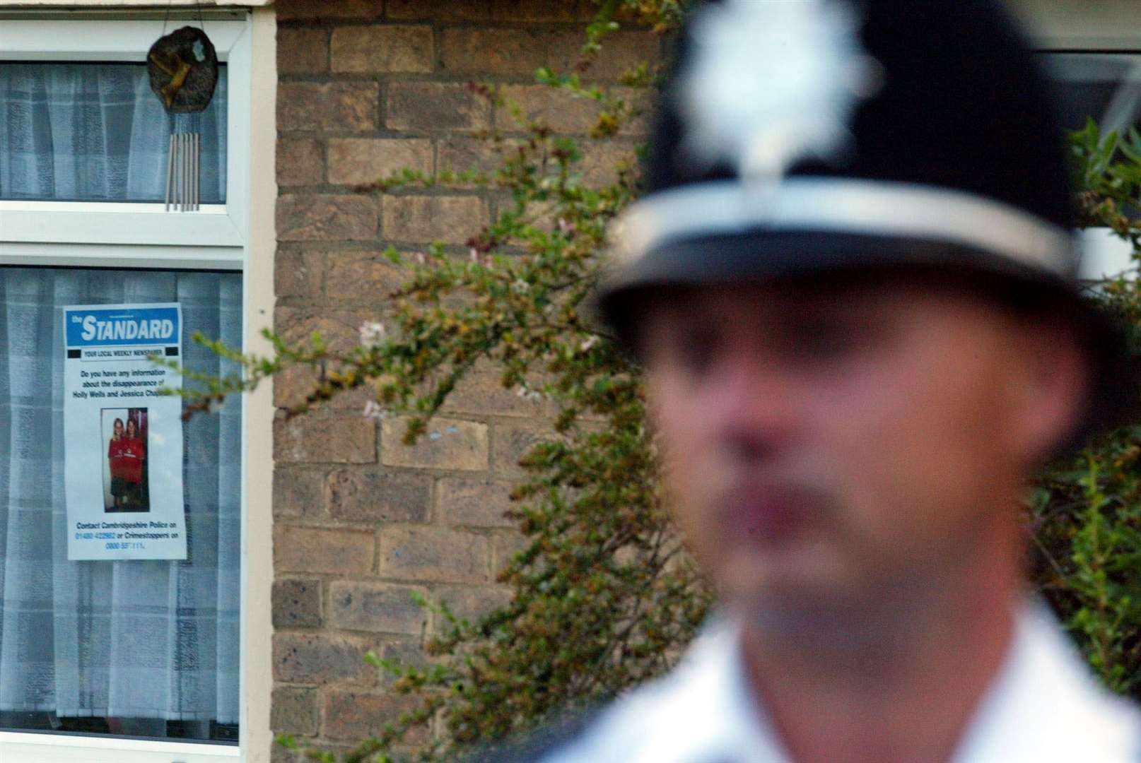 A police officer outside the home of Ian Huntley and Maxine Carr in Soham, Cambridgeshire, with the poster in the window (PA)