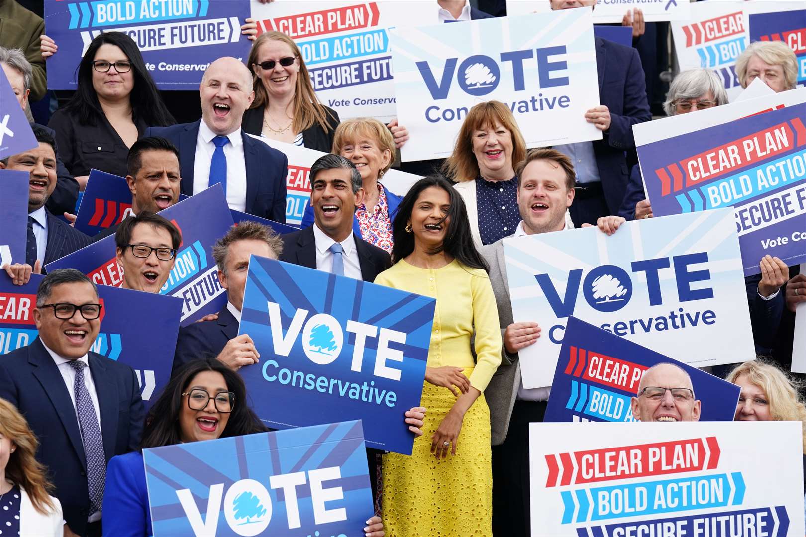 Rishi Sunak in a suit and his wife Akshata Murty in a yellow dress surrounded by campaigners holding ‘vote Conservative’ signs (James Manning/PA)