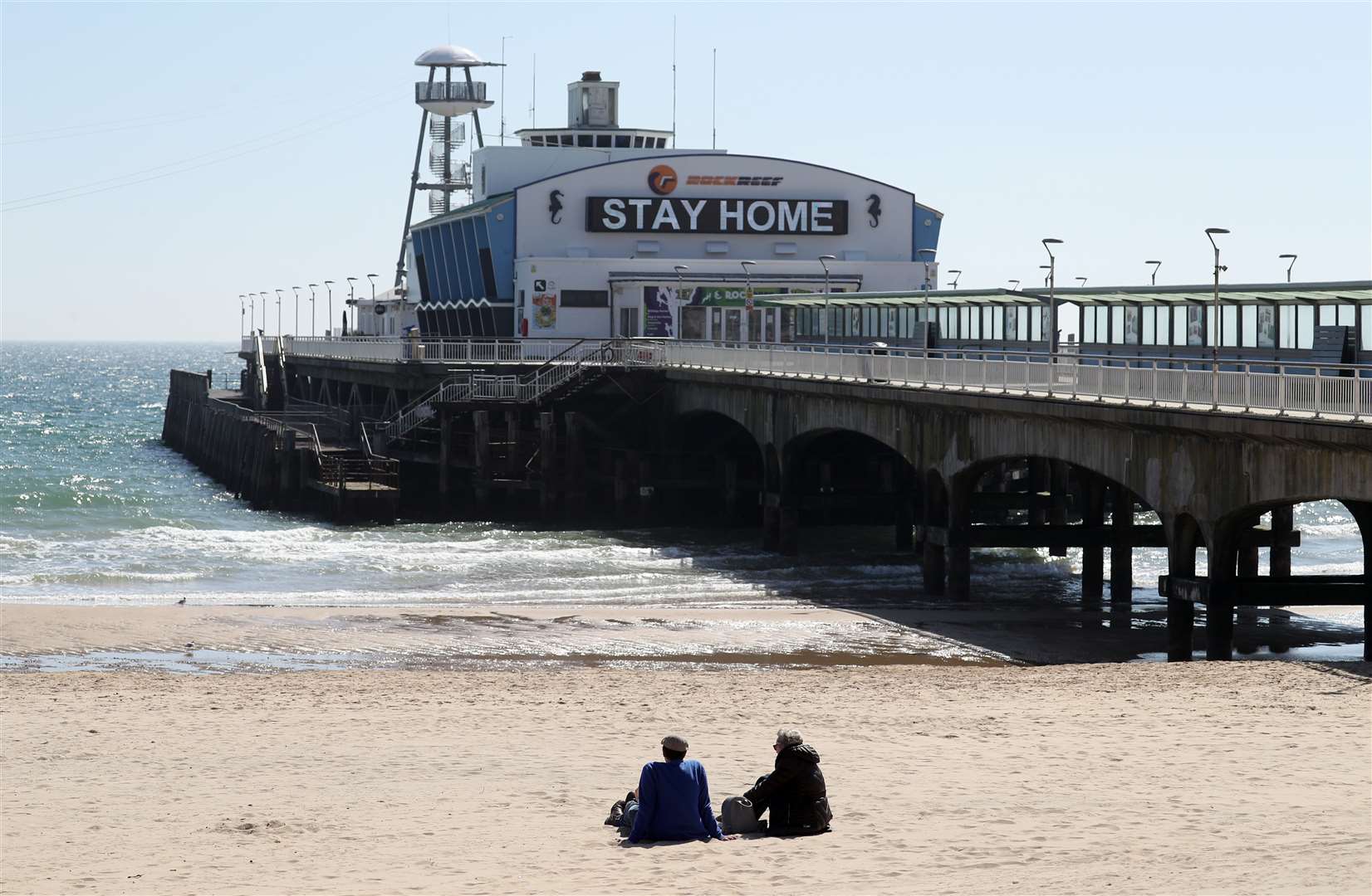People sit under a ‘Stay home’ sign on a deserted Bournemouth beach (Andrew Matthews/PA)