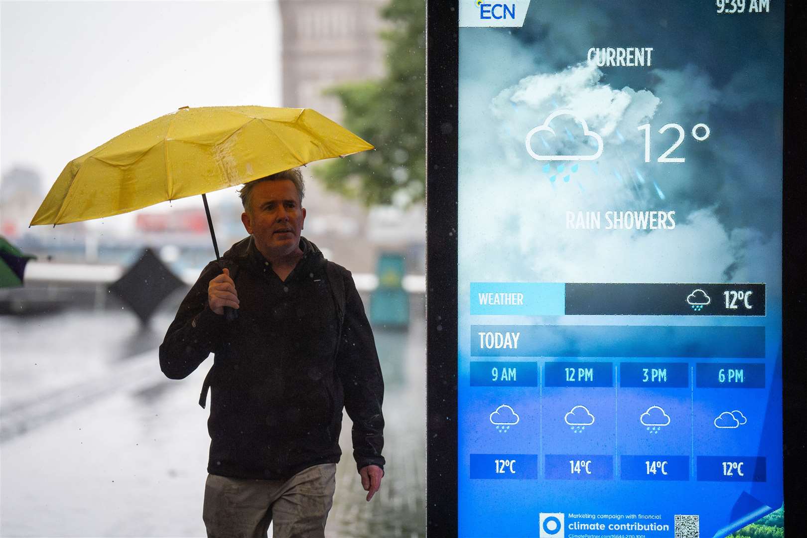 A man shelters under a yellow umbrella while a digital information board in the foreground displays a rainy weather forecast (James Manning/PA)