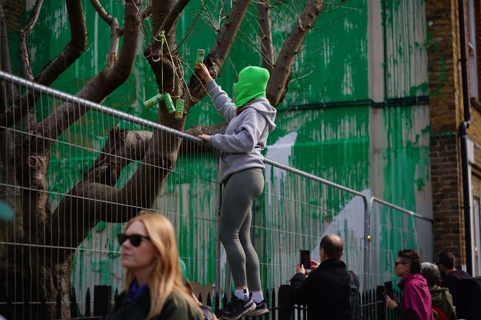 A person ties green cans to the tree in front of the Banksy artwork in Finsbury Park, London after it was defaced with white paint (Victoria Jones/PA)