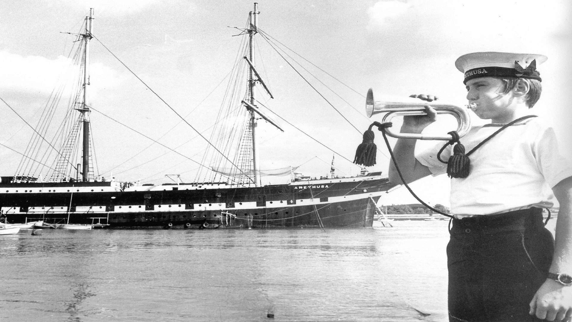 A bugle boy sounds reveille on-shore at Upnor with TS Arethusa as a backdrop.